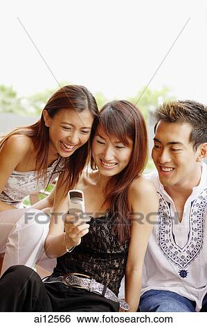 Stock Photograph - Young women and young man looking at mobile phone, smiling. Fotosearch