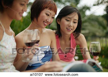 Young women browsing through magazine, wine glasses in hand View Large Photo Image Stock Photo - Young women browsing through magazine, wine glasses in hand. Fotosearch