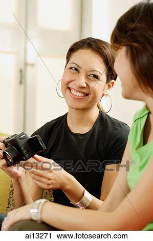 Stock Image - Young women sitting side by side, one holding camera. Fotosearch