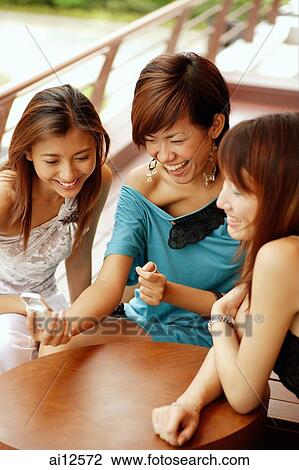 Stock Image - Young women sitting side by side, looking at mobile phone. Fotosearch