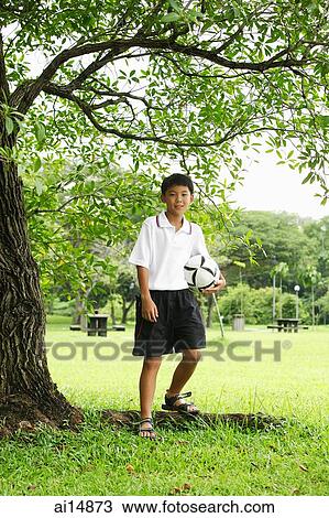 Boy holding ball, standing under tree View Large Photo Image Stock Image - Boy holding ball, standing under tree. Fotosearch