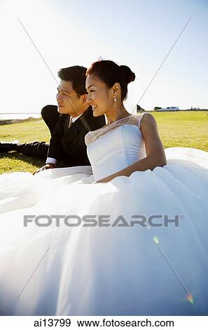 Stock Photo - Bride and groom sitting outdoors on grass looking away. Fotosearch