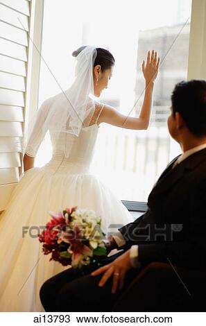 Stock Image - Bride looking out of window, groom sitting with bouquet of flowers looking at her. Fotosearch