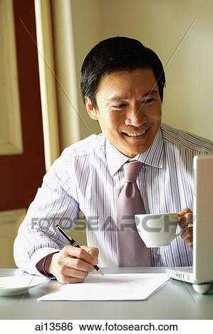 Businessman holding cup and pen, looking at laptop View Large Photo Image Stock Photograph - Businessman holding cup and pen, looking at laptop. Fotosearch
