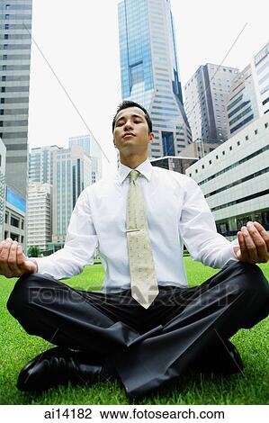 Stock Image - Businessman sitting on grass, legs crossed, buildings in the background. Fotosearch
