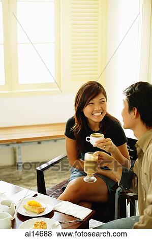 Stock Photo - Couple at cafe, holding coffee mugs, food on table. Fotosearch