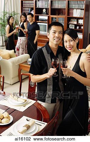 Couple at home, holding wine glasses, smiling at camera, people in the background View Large Photo Image Stock Image - Couple at home, holding wine glasses, smiling at camera, people in the background. Fotosearch