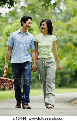 Picture - Couple holding hands and walking in park, man carrying picnic basket. Fotosearch