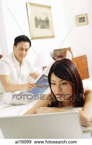 Couple in bedroom, woman lying on bed, looking at laptop, man reading newspaper behind her View Large Photo Image Stock Photo - Couple in bedroom, woman lying on bed, looking at laptop, man reading newspaper behind her. Fotosearch
