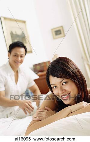 Couple in bedroom, woman lying on bed, looking at camera, man sitting behind her View Large Photo Image Stock Photography - Couple in bedroom, woman lying on bed, looking at camera, man sitting behind her. Fotosearch