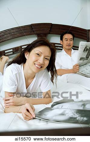 Couple in bedroom, woman smiling at camera, man reading newspaper in the background View Large Photo Image Stock Image - Couple in bedroom, woman smiling at camera, man reading newspaper in the background. Fotosearch