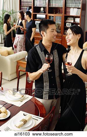 Stock Image - Couple in dining room, holding wine glasses, people in the background. Fotosearch