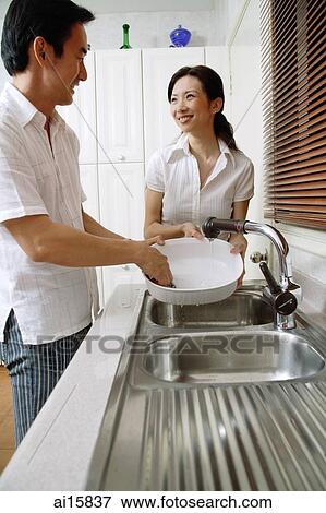 Stock Photo - Couple in kitchen, man washing dishes, woman standing next to him, smiling. Fotosearch