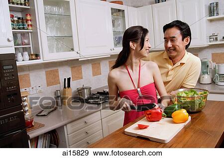 Stock Photo - Couple in kitchen, woman chopping vegetables, looking over shoulder to smile at man. Fotosearch