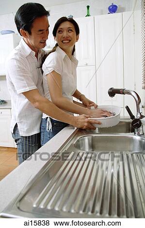 Stock Photo - Couple in kitchen, woman washing dishes, man standing behind her. Fotosearch