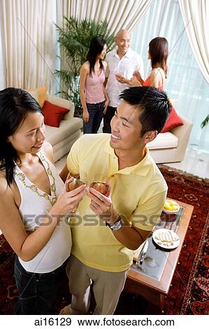 Couple in living room holding wine glasses, smiling at each other, people in the background View Large Photo Image Stock Photo - Couple in living room holding wine glasses, smiling at each other, people in the background. Fotosearch