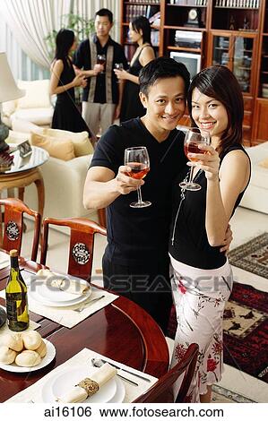 Couple in living room, raising wine glasses to camera, people in the background View Large Photo Image Stock Photograph - Couple in living room, raising wine glasses to camera, people in the background. Fotosearch