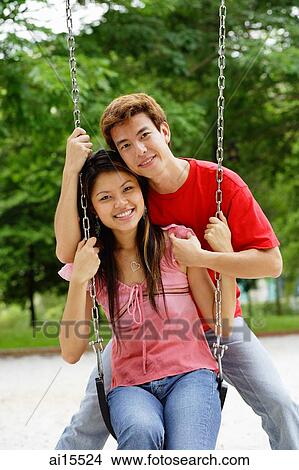 Picture - Couple in playground, woman sitting on swing, man behind her, smiling at camera. Fotosearch