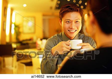 Stock Image - Couple in restaurant, man holding cup, facing woman, over the shoulder view. Fotosearch