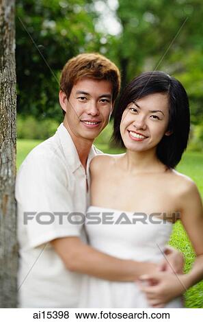 Stock Photo - Couple leaning on tree, embracing, looking at camera. Fotosearch