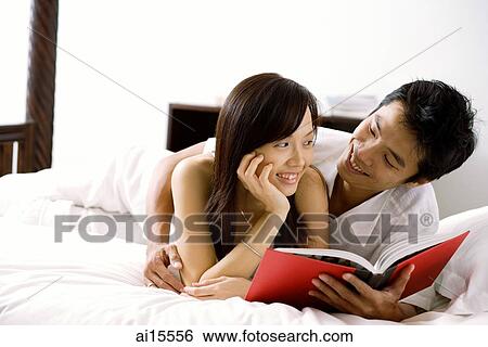 Stock Photograph - Couple lying on bed, holding book, smiling at each other. Fotosearch