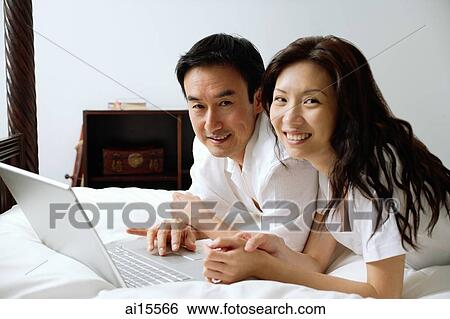 Couple lying on bed, using laptop, smiling at camera View Large Photo Image Stock Photograph - Couple lying on bed, using laptop, smiling at camera. Fotosearch