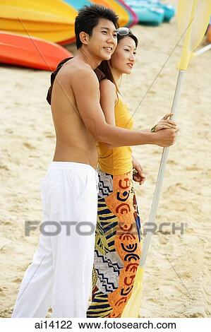 Stock Image - Couple on beach holding kayak paddle, looking away. Fotosearch