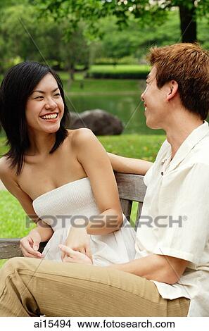 Picture - Couple sitting face to face on park bench, woman laughing. Fotosearch