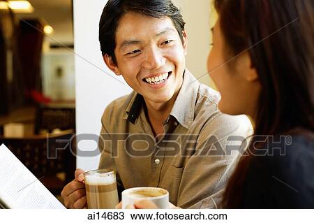 Stock Image - Couple sitting, holding coffee cups, smiling at each other. Fotosearch