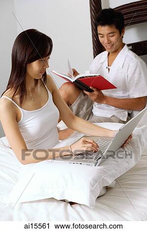 Stock Image - Couple sitting on bed, man reading book, woman using laptop. Fotosearch