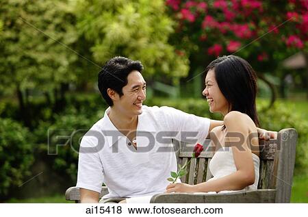 Couple sitting on park bench, looking at each other, woman holding rose View Large Photo Image Stock Photo - Couple sitting on park bench, looking at each other, woman holding rose. Fotosearch