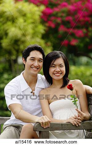 Couple sitting on park bench, looking at camera, woman holding rose View Large Photo Image Stock Photograph - Couple sitting on park bench, looking at camera, woman holding rose. Fotosearch