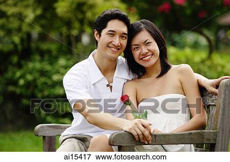 Couple sitting on park bench, smiling at camera, woman holding rose View Large Photo Image Stock Photo - Couple sitting on park bench, smiling at camera, woman holding rose. Fotosearch