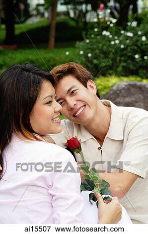 Stock Photo - Couple sitting on park bench, woman holding a rose, looking away. Fotosearch