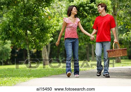 Stock Image - Couple walking in park, looking at each other, man carrying picnic basket. Fotosearch