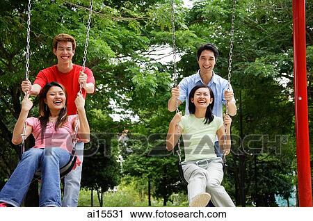 Stock Image - Couples in playground, women on swings, men pushing them. Fotosearch