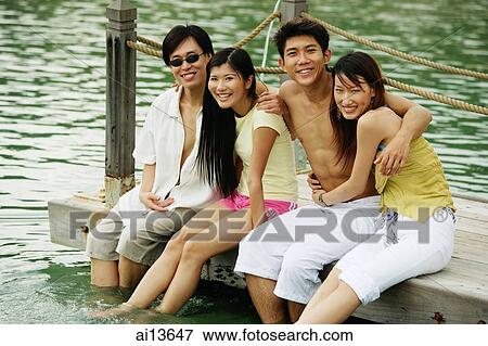 Stock Photo - Couples sitting side by side on jetty, arms around each other. Fotosearch
