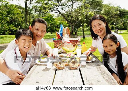 Stock Image - Family sitting at picnic table, looking at camera. Fotosearch