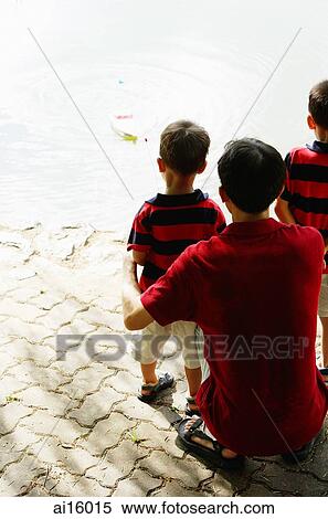 Stock Photography - Father and two sons looking at toy boat on lake. Fotosearch
