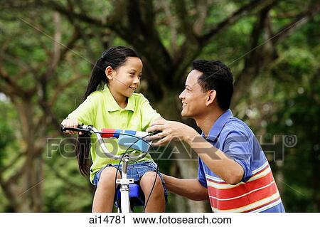 Stock Image - Girl on bicycle, father crouching next to her. Fotosearch