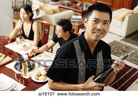 Stock Photo - Man holding wine bottle, looking at camera, people in the background, sitting around dining table. Fotosearch