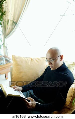 Stock Image - Man in black turtleneck, sitting on sofa in living room, reading book. Fotosearch
