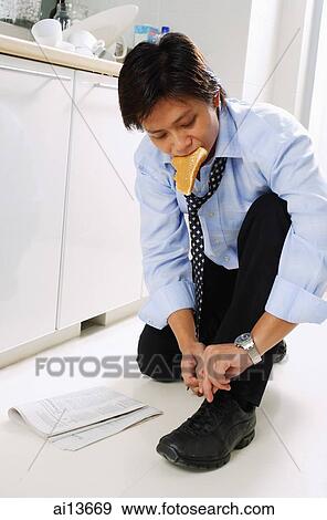 Stock Photo - Man in kitchen, tying shoelace, slice of bread in mouth. Fotosearch