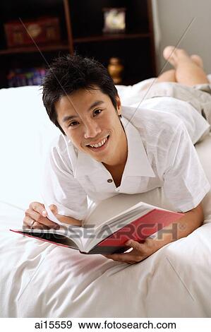Man lying on bed, holding book, smiling at camera View Large Photo Image Stock Photo - Man lying on bed, holding book, smiling at camera. Fotosearch