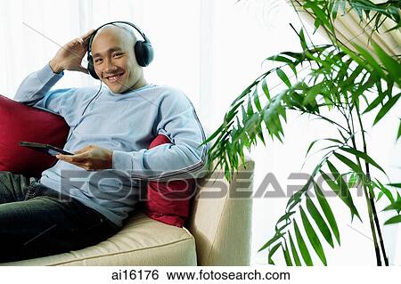 Stock Photograph - Man on sofa with headphones, listening to music, smiling at camera. Fotosearch