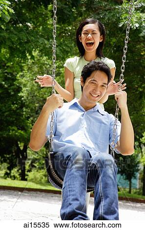 Stock Photography - Man sitting on swing, woman standing behind him, laughing. Fotosearch