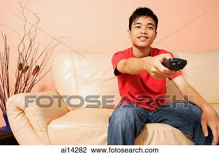 Teenage boy sitting on sofa, holding remote control, pointing at camera View Large Photo Image Stock Image - Teenage boy sitting on sofa, holding remote control, pointing at camera. Fotosearch