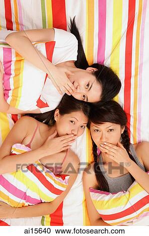 Three young women lying on bed, looking up at camera, hands over mouths View Large Photo Image Stock Image - Three young women lying on bed, looking up at camera, hands over mouths. Fotosearch