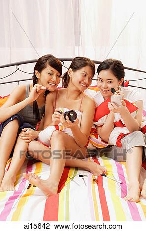 Three young women sitting on bed, side by side, applying make-up View Large Photo Image Stock Image - Three young women sitting on bed, side by side, applying make-up. Fotosearch