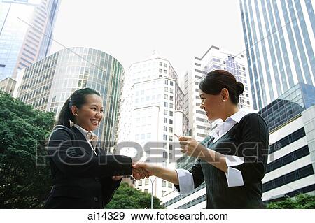 Two business women shaking hands, one holding business card, low angle view View Large Photo Image Stock Photo - Two business women shaking hands, one holding business card, low angle view. Fotosearch